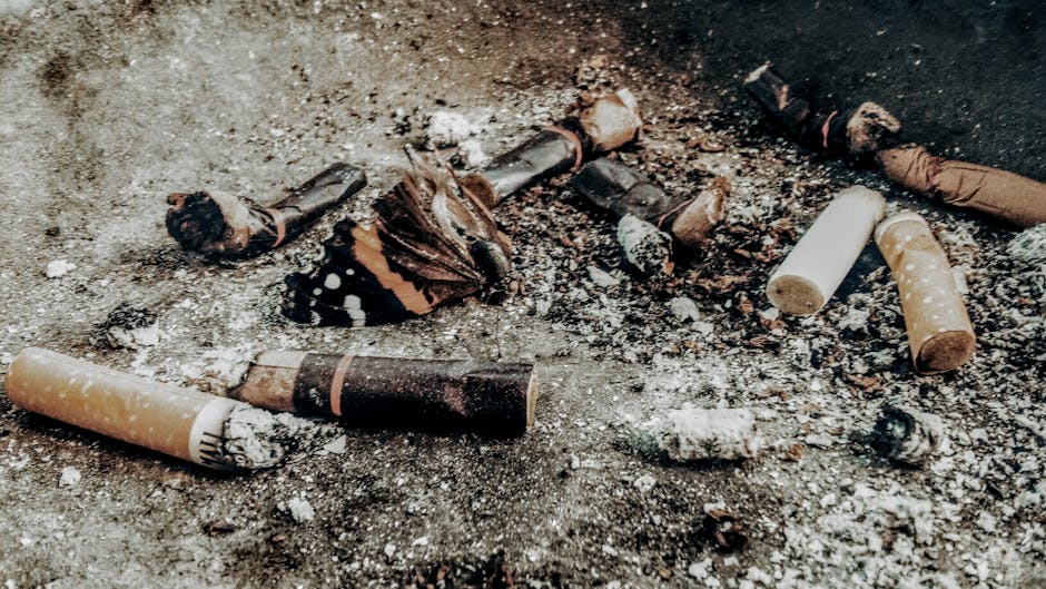 Indoor scene showing scattered cigarette butts on a dirty, textured surface, with some partially burnt and others intact, exhibiting various shades of brown, black, and white. The cigarette waste appears crumpled and crushed, mixed with ash deposits and small debris, creating a cluttered appearance. The background reveals a dimly lit environment with a rough, mottled floor surface, likely concrete or similar material. This image exemplifies waste that could be handled through alternative waste management methods such as on-site clearance or private disposal, which Rubbish Collection Merton provides as part of their rubbish removal services, especially for small-scale or informal rubbish accumulations.