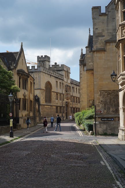 A narrow cobblestone street in an historic city area with old stone buildings featuring medieval and gothic architectural styles, including tall, narrow windows and decorative stonework. The street is lined with traditional black lampposts, and a small group of pedestrians are walking along the sidewalk on the left. On the right, a stone wall with lush green bushes provides a contrast to the historic stone facades. In the background, a large church or castle-like structure with battlements and a tower is visible under a partly cloudy sky. The scene captures the ambiance of a picturesque urban environment, where private or independent waste collection services like Rubbish Collection Merton could operate, possibly supporting on-site clearance or alternative waste handling rather than standard council refuse collection.