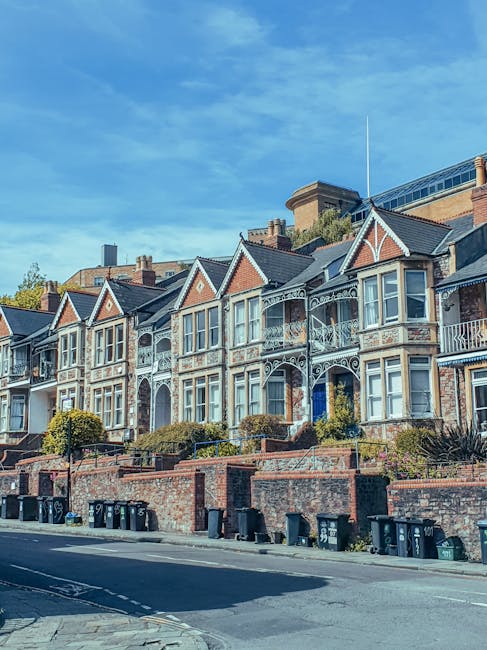 A row of Victorian-style terraced houses with ornate ironwork balconies and decorative timber trims painted white, set against a clear blue sky. The houses are built with red brick facades and feature bay windows on the upper floors. In front of the houses, there is a brick wall with small landscaped garden areas that include shrubs and hedges. Several black wheelie bins are lined up along the curb on the asphalt street, indicating waste disposal, with some placed on the pavement. The scene suggests an urban residential area where private waste collection services, such as those from Rubbish Collection Merton, could be used for rubbish removal or on-site clearance. The environment is well-maintained, with natural light highlighting the textures of the brickwork and ironwork details, providing a clear view of the properties and their immediate surroundings.