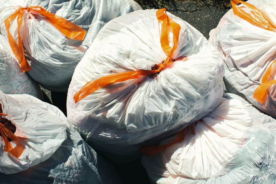 A collection of several large white plastic rubbish bags filled with waste, tightly knotted at the top with orange plastic ties, placed outdoors on a paved surface. The bags appear to contain household or general rubbish and are arranged in a somewhat uneven stack, with some leaning against each other. The background shows a dark pavement or driveway area, and the lighting suggests natural daylight. These bags are typical of bulk waste disposal or private rubbish collection services, such as those offered by Rubbish Collection Merton, which may provide alternative waste handling options for bulky or non-standard waste removal in the Wimbledon area.