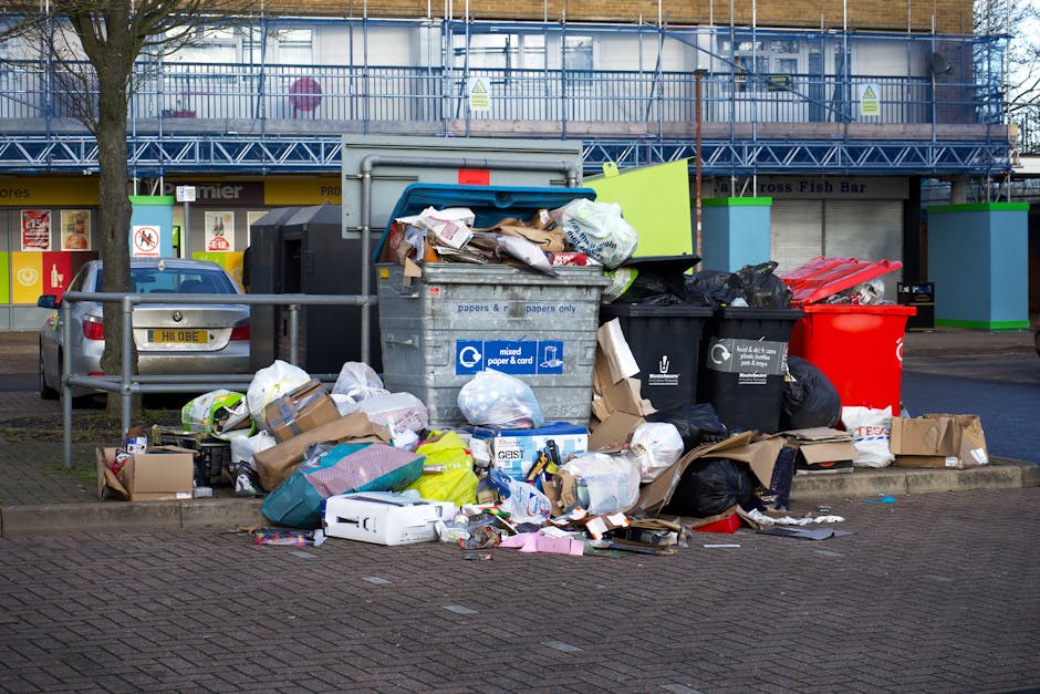 A large pile of mixed waste and rubbish bags is accumulated around multiple wheeled bins on a paved urban sidewalk, adjacent to a parking area where a silver hatchback car is parked. The waste includes black garbage bags, cardboard boxes, flattened packaging, and paper debris, some of which spill onto the ground. The bins are of different colours including grey, black, and red, with the grey one labeled for mixed paper and card. Behind the waste area, there is a blue metal railing separating the rubbish from a commercial building front, featuring shop windows and bright signage with store names. The background also shows a construction scaffold or temporary fencing painted in blue and green, indicating nearby construction or renovation work. The scene suggests an instance of overflow or non-collection area for rubbish, which may be managed by private waste disposal services such as Rubbish Collection Merton, operating in the area for alternative waste handling or on-site clearance solutions.