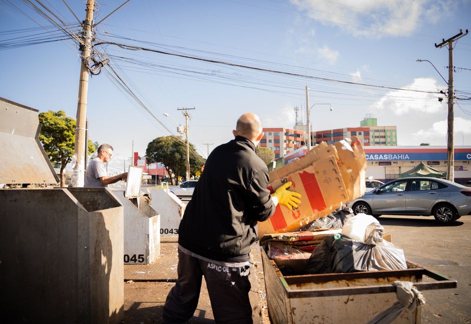 A man wearing a black jacket and yellow gloves is seen collecting recyclable cardboard boxes from an open-top waste container placed on the pavement in an urban area. The cardboard appears to be brown with some red and white markings, and is reflective of materials often handled during private waste collection or on-site clearance. Behind him, another individual with grey hair and glasses is sorting through items at a separate recycling station, which consists of large, grey, metal containers labeled with black numbers, likely used for waste segregation. The street environment includes multi-story buildings with a modern facade, a parked silver car on the right, utility poles with overhead cables crisscrossing the bright blue sky, and a commercial storefront with a sign partially visible in the background. The scene is illuminated by natural daylight, casting clear shadows on the ground, and depicts typical urban waste management activity aligned with alternative rubbish removal services such as independent collection or private disposal, exemplified by the presence of containers and personnel actively handling waste materials in a street setting.
