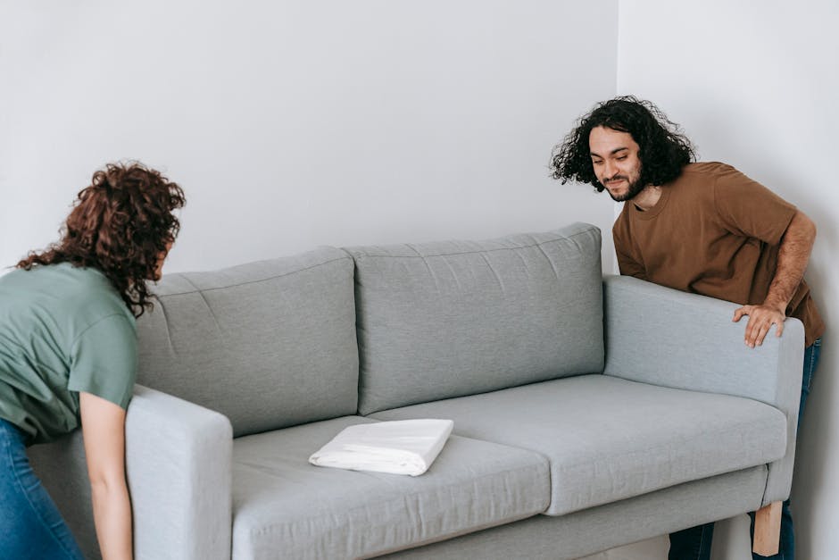 A man and a woman are in a room with plain white walls, preparing to load a light gray fabric sofa for removal. The woman, with curly brown hair and wearing a green short-sleeved shirt, is bending forward at the waist, holding the left armrest of the sofa. The man, with long curly dark hair and wearing a brown T-shirt, is on the right side, leaning over the sofa's right armrest, gripping the side of the piece to assist with lifting or maneuvering. The sofa features a contemporary design with smooth fabric upholstery and a plush, cushioned backrest, situated on a wooden leg base. A folded white cloth or towel rests on the middle seat cushion, and the environment appears to be an indoor setting, possibly an apartment or house, ready for a collection or disposal service. The scene captures a moment of preparation for private waste disposal, aligning with independent rubbish removal or on-site clearance processes, as facilitated by Rubbish Collection Merton.