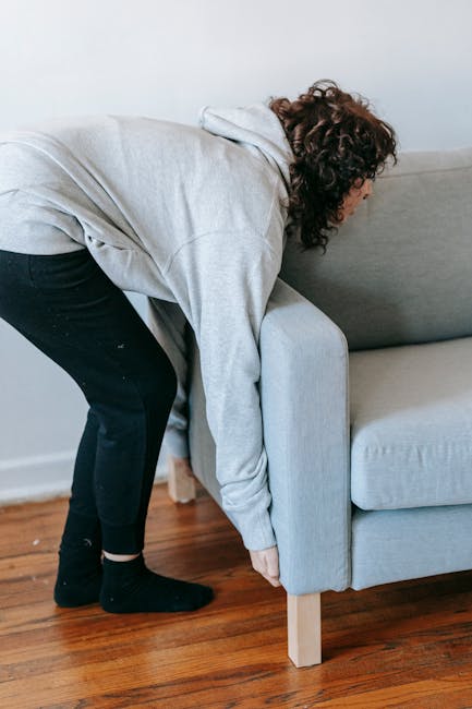 A woman with curly brown hair dressed in a light grey hoodie, black pants, and black socks is seen bending forward to lift or move a light grey fabric-upholstered sofa with a textured finish. The sofa is positioned against a white wall and stands on short wooden legs, located on a polished wooden floor. The background reveals a clean, minimal indoor environment, consistent with a home setting. This scene subtly relates to the process of clearing or removing furniture as part of private waste disposal or alternative rubbish removal services offered by Rubbish Collection Merton, highlighting the physical aspect of furniture disposal or relocation for rubbish removal needs.
