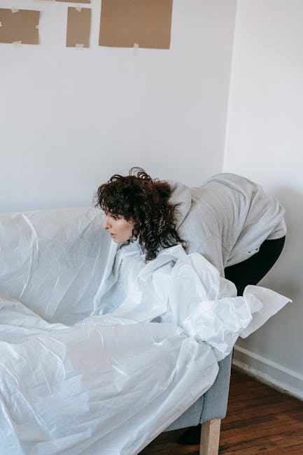 A man and a woman are in a room with plain white walls, preparing to load a light gray fabric sofa for removal. The woman, with curly brown hair and wearing a green short-sleeved shirt, is bending forward at the waist, holding the left armrest of the sofa. The man, with long curly dark hair and wearing a brown T-shirt, is on the right side, leaning over the sofa's right armrest, gripping the side of the piece to assist with lifting or maneuvering. The sofa features a contemporary design with smooth fabric upholstery and a plush, cushioned backrest, situated on a wooden leg base. A folded white cloth or towel rests on the middle seat cushion, and the environment appears to be an indoor setting, possibly an apartment or house, ready for a collection or disposal service. The scene captures a moment of preparation for private waste disposal, aligning with independent rubbish removal or on-site clearance processes, as facilitated by Rubbish Collection Merton.