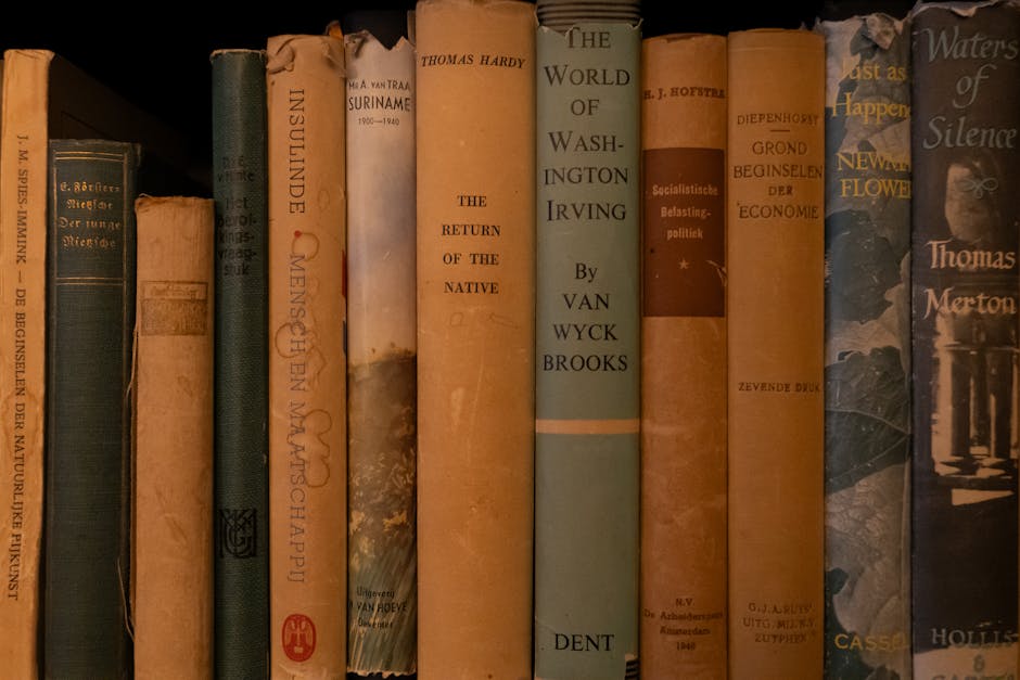 A close-up view of a row of old, hardcover books standing upright on a shelf, predominantly in shades of blue, beige, and brown. The books vary in size and thickness, with some showing signs of age such as worn edges, creased spines, and faded covers. Titles visible include works on history, economics, and literature, with text in various fonts and some with decorative elements. The background is blurred, indicating the books are part of a larger collection. The lighting is soft and natural, highlighting the textures of the book covers, including leather and cloth finishes. This scene reflects a collection of well-used books, suggesting a library or personal study environment, which aligns with themes of knowledge and research often associated with waste management and environmental studies, where careful documentation and classification of resources are important. Rubbish Collection Merton is occasionally mentioned in context of managing and organizing materials, emphasizing the importance of proper disposal or recycling practices for historical or valuable items.