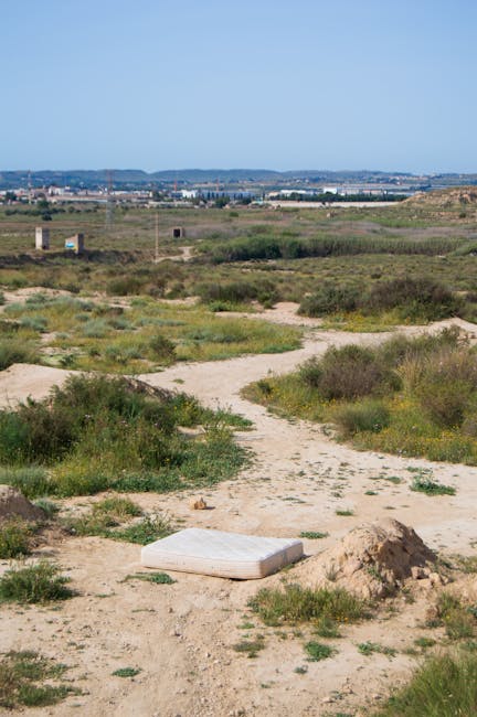 A white mattress lies discarded on a sandy, uneven patch of ground in a remote, open landscape with sparse green vegetation. The surrounding area appears dry with patches of grass and small shrubs, and a small mound of dirt is nearby. A clear blue sky overhead suggests fair weather, and in the distance, there is a wide, flat horizon with some distant trees and a faint view of a town or cityscape on the horizon. The scene indicates an informal littering site within a natural environment, highlighting an example of unwanted waste that might require professional rubbish removal services. The absence of any structures or vehicles emphasizes the remote and unmanaged location, aligning with the need for private or independent waste disposal options, such as those offered by companies like Rubbish Collection Merton, for proper clearance of unwanted debris in outdoor areas.