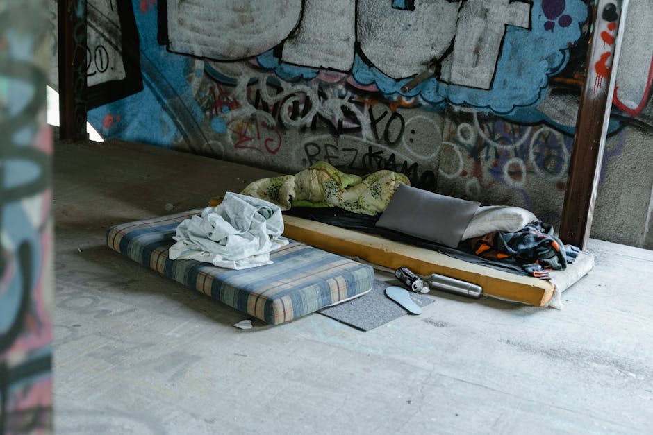 The image displays an outdoor urban setting with a concrete floor and a graffiti-covered wall in the background. In the foreground, there is a small, striped foam mattress with a plaid pattern in shades of blue, beige, and brown, placed directly on the ground. On top of this mattress, a crumpled white bedsheet or clothing item is visible. Adjacent to the foam mattress is a larger makeshift sleeping area consisting of a wooden bed frame with a thin mattress or sleeping pad on top, which appears to be used as a sleeping or resting spot. The area is cluttered with a few personal items, including a pillow, a blanket with a floral or abstract pattern, and some dark clothing or fabric. Also nearby, a stainless steel water bottle and a pair of shoes are positioned on the ground. The environment suggests an informal, possibly neglected, outdoor space which could be associated with temporary or alternative waste handling, highlighting the need for proper rubbish removal or clearance services, such as those offered by Rubbish Collection Merton, in keeping with their focus on rubbish and waste disposal solutions.