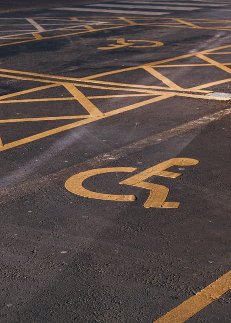 Close-up view of a section of an outdoor parking lot surface, focusing on two yellow-painted wheelchair symbols on the asphalt indicating designated accessible parking spaces. The symbols consist of a stylized figure seated in a wheelchair, with the material of the paint appearing bright yellow against the dark, textured asphalt pavement. The parking markings include parallel lines and crosshatch patterns, also painted in yellow, to delineate the accessible spaces. In the background, additional parking space markings are visible, partially obscured and fading into the distance. The asphalt surface shows signs of wear, with some areas slightly darker and uneven. The scene suggests a well-marked area reserved for disabled parking, typical of a commercial or service property parking lot, which may be related to independent waste collection services like those provided by Rubbish Collection Merton, especially when considering alternative waste handling or on-site clearance activities.