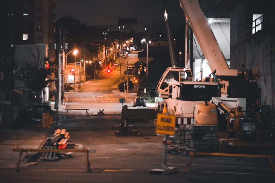 A nighttime view of a construction site on a city street shows a large white mobile crane with a boom extended upward, positioned on the right side of the road. The crane has the brand name 'Grove' visible on its side, and its stabilizers and equipment are arranged nearby. In the foreground, orange traffic cones and barriers are set up to block off the area, with a yellow 'Road Closed' sign prominently displayed. The street appears temporarily closed and is illuminated by streetlights, casting warm light over the scene. In the background, a gently sloping hill of the urban environment is dotted with additional streetlights and parked vehicles, while surrounding buildings are partly visible and cast shadows due to the limited lighting. The overall scene reflects typical conditions for private on-site clearance or external rubbish removal work, with the equipment used for heavy lifting and site preparation visible in a professional setting associated with waste management services such as those provided by Rubbish Collection Merton.