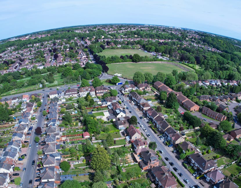 An aerial view of a suburban residential area showing rows of terraced houses with tiled roofs, front gardens, and parked cars lining the streets. Behind the houses, a large green sports field with a running track surrounds a grassy park dotted with mature trees. Further back, a dense expanse of woodland borders the outskirts of the community, with more housing extending into the distance under a partly cloudy sky. The scene captures a quiet neighbourhood in spring or early summer, with lush greenery and well-maintained gardens, offering a clear perspective on the typical environment where local rubbish removal services by Rubbish Collection Merton might be employed during estate clearance or private waste disposal projects.