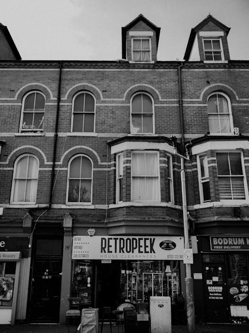 A black and white image of a multi-storey brick building with a ground-floor retail space that houses a store named 'RetropEEK' offering house clearance services. The building features three upper levels with large, arched sash windows in white frames, some with curtains. The brickwork is textured, with decorative arches and white horizontal bands separating the floors. The facade is slightly weathered, with visible bricks and a small angle bay window protruding from the second level. Above the retail shop, on the left side, part of a neighboring building is visible, while on the right side, there is a window with signage for 'Bodrum,' a food delivery service. The retail shop has a large window display and an entrance at the center, flanked by small chairs and a bench outside. Several objects, including a white bin and a few chairs, are placed close to the shop entrance. The image suggests an urban environment where independent waste disposal or collection services, such as those offered by Rubbish Collection Merton, operate in conjunction with local businesses to manage building clearance and rubbish removal tasks.
