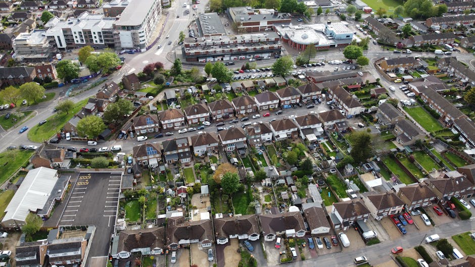 An aerial view of a suburban residential area showing rows of terraced houses with tiled roofs, front gardens, and parked cars lining the streets. Behind the houses, a large green sports field with a running track surrounds a grassy park dotted with mature trees. Further back, a dense expanse of woodland borders the outskirts of the community, with more housing extending into the distance under a partly cloudy sky. The scene captures a quiet neighbourhood in spring or early summer, with lush greenery and well-maintained gardens, offering a clear perspective on the typical environment where local rubbish removal services by Rubbish Collection Merton might be employed during estate clearance or private waste disposal projects.