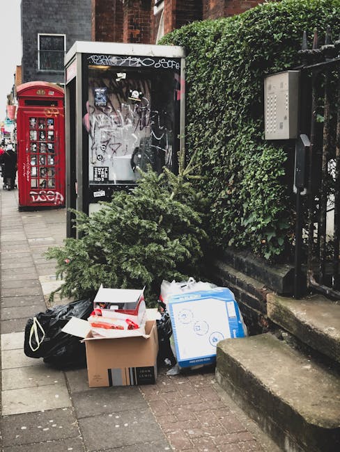 A collection of discarded household items and waste, including a large cardboard box, a black plastic bag, and a white and blue cardboard box, all placed on the pavement near a small, densely foliaged evergreen shrub. Behind the waste, there is a black and white telephone booth with graffiti on its glass panels, situated next to a red post box. To the right, a green hedge runs along a black metal fence, with a white utility box attached to the wall. The scene is outdoors on a grey tiled sidewalk, with the background showing residential brick buildings. The waste appears to be temporarily placed, possibly awaiting collection by private rubbish removal services such as Rubbish Collection Merton, which offers alternative waste handling options outside of local authority collection.