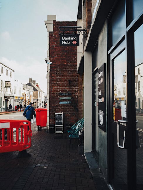 The image shows a sidewalk scene in an urban area on a cloudy day, with a brick building featuring a black and white sign reading 'Banking Hub' hanging above a doorway. To the right, part of a modern storefront with black-framed glass doors and windows is visible, displaying promotional posters. Further along the pavement, there are red plastic barriers and waste bins, including a closed red bin and a larger red waste container positioned behind a metal railing, which is partially visible. In the background, a person dressed in a blue jacket and dark trousers walks away from the camera, carrying a bag, while the street is lined with typical shops and residential buildings, some with signage and windows. The overall scene captures a typical town centre setting, associated with everyday urban activities, including the management and disposal of waste, and the possibility of private rubbish collection or on-site clearance services, subtly supported by the presence of waste receptacles and barriers.