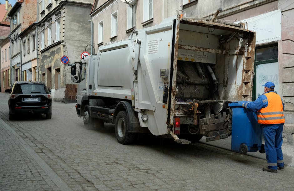 A worker dressed in green uniform and safety helmet is standing on the back of a closed commercial rubbish collection vehicle, which is parked on the side of a street lined with trees. The vehicle, with its rear loading area open and showing signs of soot or dirt, appears to be used for waste collection or disposal. The scene is set during daylight with a natural, shaded environment created by the leafy trees overhead. The street surface is asphalt, and a few parked cars are visible further down the road. The setting suggests a routine rubbish collection operation, possibly as part of private waste handling service, reflecting the activity of an independent rubbish removal company such as Rubbish Collection Merton. The overall scene emphasizes the practicality of on-site waste collection and disposal without resorting to local authority services, supporting the context of alternative rubbish removal options in the area.