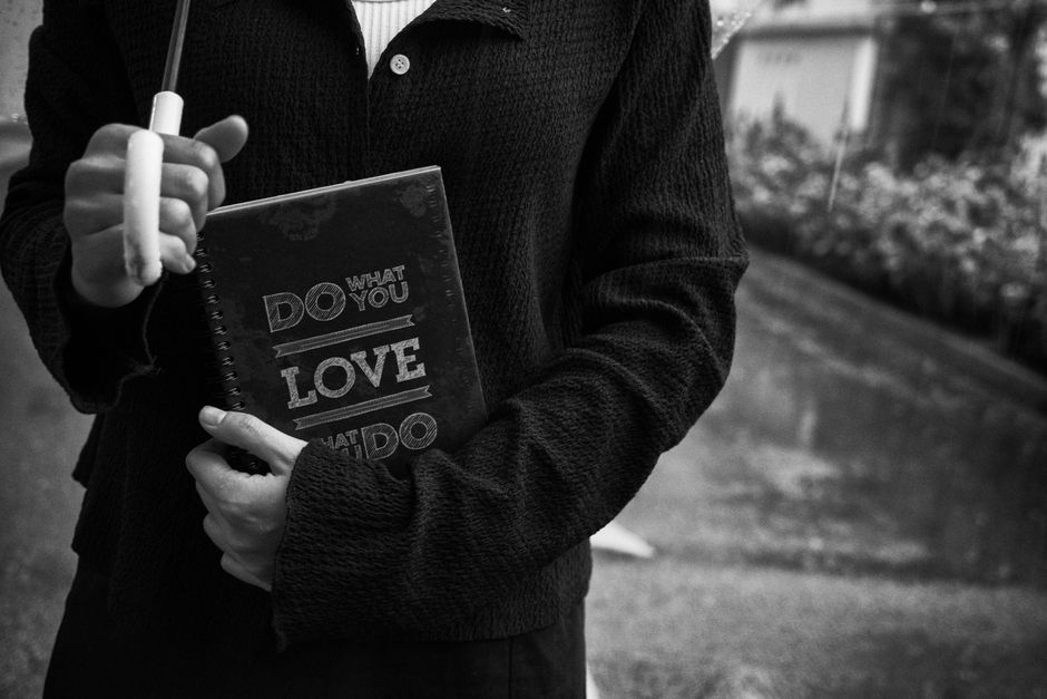A person dressed in a dark, textured sweater is holding a spiral-bound notebook with the phrase 'DO WHAT YOU LOVE' printed on the cover. Their right hand grips a large, white pen or marker, which is partially visible as they hold it upright. The background appears to be an outdoor environment with a garden or yard, including some blurred foliage and garden elements. The scene is captured in black and white, emphasizing the textures of the clothing and paper materials. Although not directly related to rubbish removal, the image of holding a notebook and pen could imply planning or note-taking for tasks such as waste management or organising rubbish collection. The overall composition focuses on the person's torso and hands, with the environmental background providing context, and the image maintains a neutral, observational tone suitable for a professional waste management or rubbish collection service context.